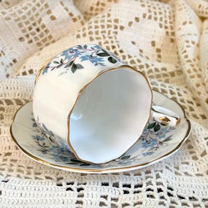May include: A white porcelain teacup and saucer set featuring a delicate pattern of light blue flowers and green leaves. The rim of both the cup and the saucer is finished with a thin gold trim, and the cup has a scalloped edge design.