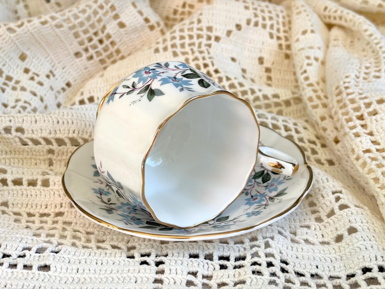 May include: A white porcelain teacup and saucer set featuring a delicate pattern of light blue flowers and green leaves. The rim of both the cup and the saucer is finished with a thin gold trim, and the cup has a scalloped edge design.
