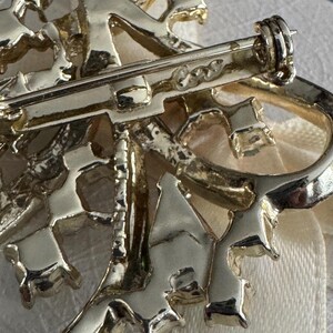 May include: Close-up of a gold-toned brooch with an intricate, openwork design. The brooch features a floral or abstract motif with a pin closure on the back. The metal has a shiny finish, and the overall style suggests a vintage or antique piece.