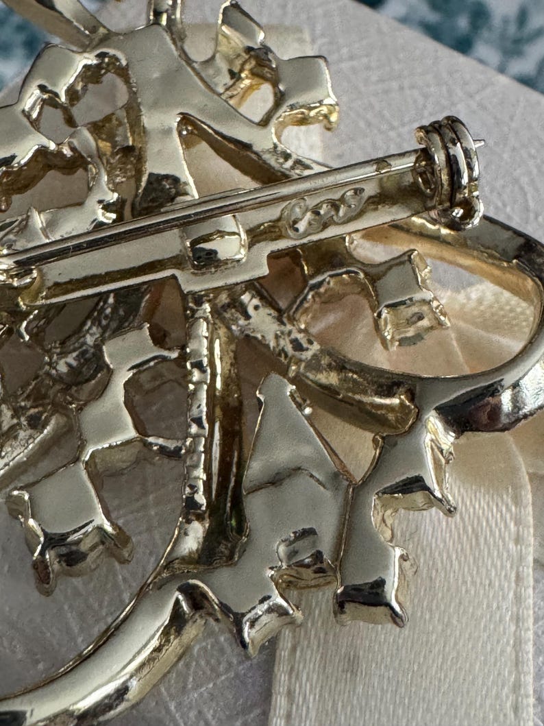 May include: Close-up of a gold-toned brooch with an intricate, openwork design. The brooch features a floral or abstract motif with a pin closure on the back. The metal has a shiny finish, and the overall style suggests a vintage or antique piece.