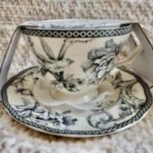 May include: A cream-colored ceramic teacup and saucer set featuring a detailed black and white toile-style print of birds and floral patterns. The rim of both the cup and saucer is decorated with a distinct black and white circular chain border.