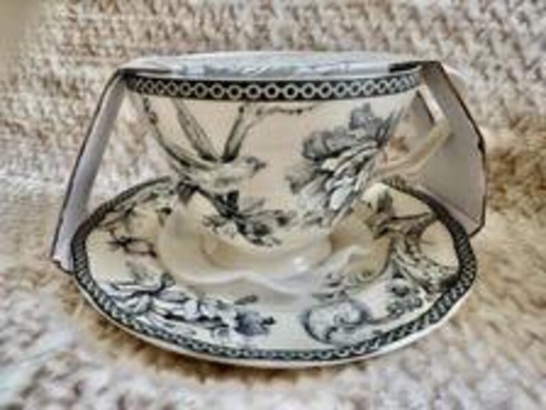 May include: A cream-colored ceramic teacup and saucer set featuring a detailed black and white toile-style print of birds and floral patterns. The rim of both the cup and saucer is decorated with a distinct black and white circular chain border.
