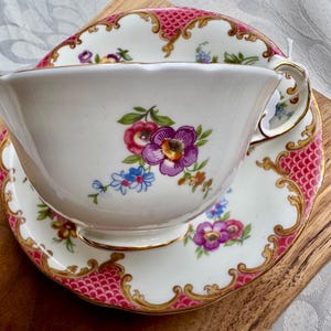 May include: A white teacup and saucer set with a floral design featuring purple, pink, and blue flowers. The rim and edges are accented with gold trim and a pink patterned border. The set is placed on a wooden surface.