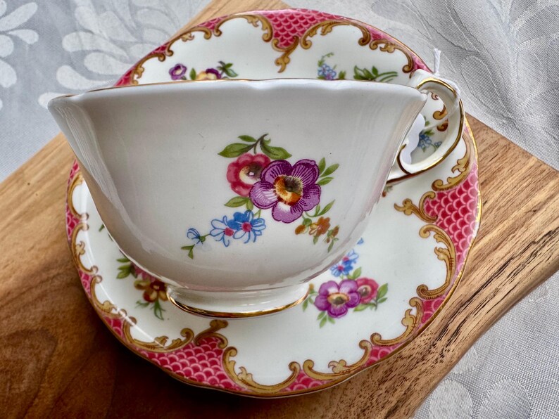 May include: A white teacup and saucer set with a floral design featuring purple, pink, and blue flowers. The rim and edges are accented with gold trim and a pink patterned border. The set is placed on a wooden surface.