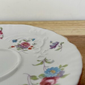 May include: White porcelain saucer with a scalloped edge and a gold rim. The saucer is decorated with embossed floral designs and colorful flowers in shades of pink, blue, and purple.