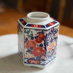 Peut inclure: Un vase hexagonal en c&eacute;ramique avec une base et un bord blancs. Le vase est d&eacute;cor&eacute; de motifs floraux rouges et oranges, d'accents bleus et de feuillage vert. Le design rappelle l'art japonais traditionnel.