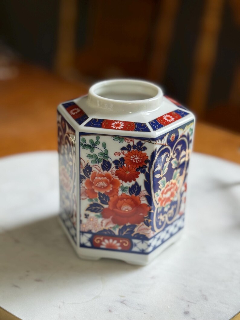 Peut inclure: Un vase hexagonal en c&eacute;ramique avec une base et un bord blancs. Le vase est d&eacute;cor&eacute; de motifs floraux rouges et oranges, d'accents bleus et de feuillage vert. Le design rappelle l'art japonais traditionnel.