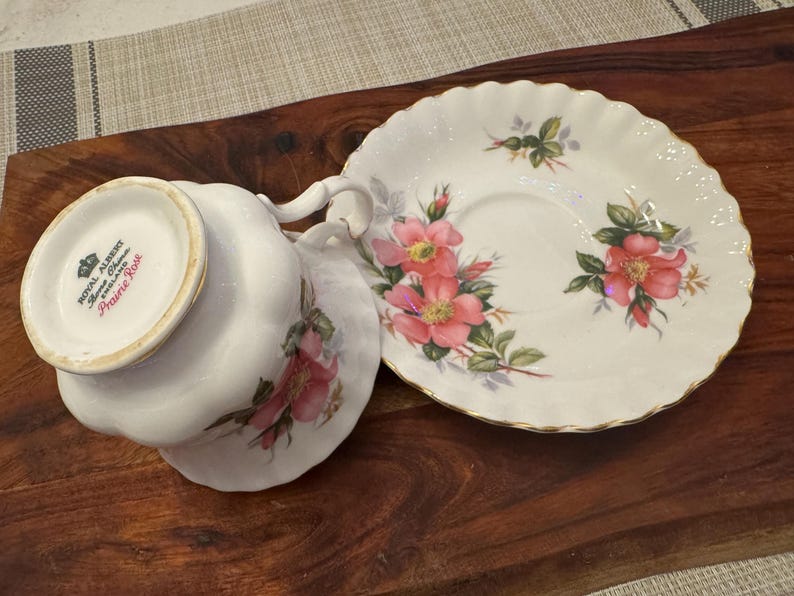 Puede incluir: Un juego de taza y platillo de t&eacute; blanco con un dise&ntilde;o floral. La taza de t&eacute; tiene un borde dorado y las palabras "Royal Albert, Bone China, England, Prairie Rose" en la parte inferior. El platillo tiene un borde festoneado y un dise&ntilde;o floral con rosas rosas.