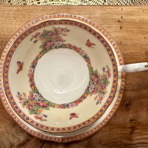 May include: A white teacup and saucer with a floral pattern and gold trim. The pattern features butterflies and flowers in shades of pink, blue, and green.