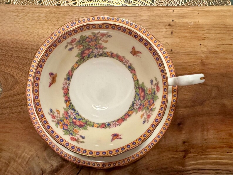 May include: A white teacup and saucer with a floral pattern and gold trim. The pattern features butterflies and flowers in shades of pink, blue, and green.