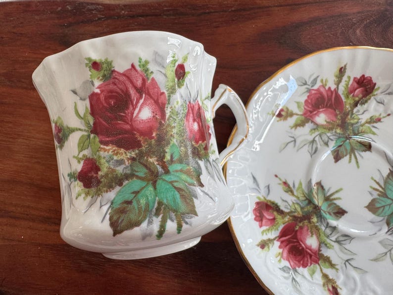 May include: A white teacup and saucer set with a floral design featuring red roses and green leaves.
