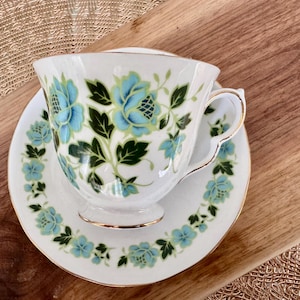 May include: A white teacup and saucer with a blue and green floral pattern. The cup has a gold rim and the saucer has a gold band around the edge.