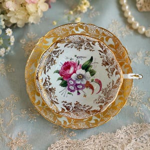 May include: A porcelain teacup and saucer set featuring a white base with a central floral bouquet of pink, purple, and white flowers. The edges are decorated with intricate gold filigree patterns and scalloped rims, with a small gold-trimmed handle.