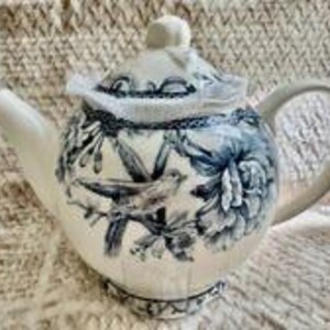 May include: A ceramic teapot featuring a cream-colored base with a dark blue transferware pattern depicting birds perched on branches and large floral blooms. The teapot includes a matching lid with a knob handle, a curved spout, and a handle.