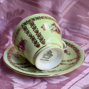 May include: A light green teacup and saucer set featuring gold leaf-patterned vertical stripes and floral bouquets. The base of the teacup is marked with the Aynsley England Bone China logo. The set has gold trim along the edges.