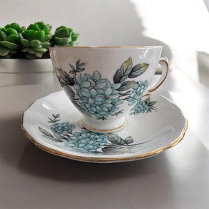 May include: A white teacup and saucer with a blue and green floral pattern. The cup and saucer have a gold trim.