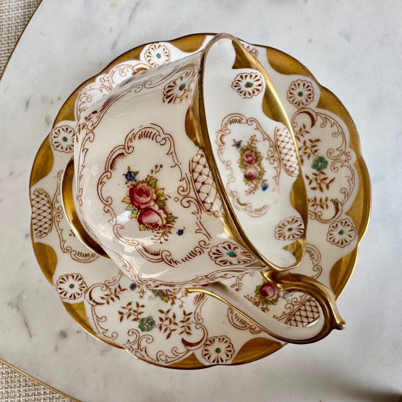 K&ouml;nnte beinhalten: Ein antikes Tee-Set, bestehend aus Tasse und Untertasse. Das wei&szlig;e Porzellan ist mit floralen Mustern in Rosa, Rot und Blau verziert und mit Gold verziert. Die Tasse ist geneigt und zeigt die detaillierten Muster.