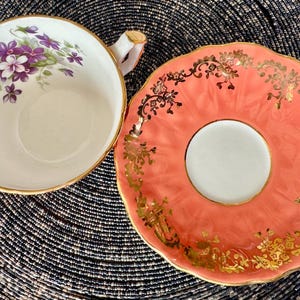 May include: A vintage teacup and saucer set with a delicate floral design. The teacup features a white background with purple and green floral accents. The saucer is a vibrant coral color with gold accents.