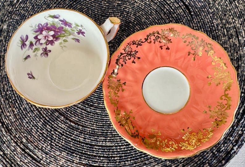 May include: A vintage teacup and saucer set with a delicate floral design. The teacup features a white background with purple and green floral accents. The saucer is a vibrant coral color with gold accents.