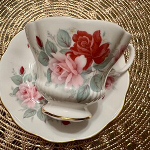 May include: A white teacup and saucer with a floral pattern featuring pink and red roses. The cup and saucer have a gold trim.