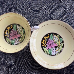 May include: A set of two yellow teacups with floral designs. The cups feature black outlines and pink, yellow, and green flowers. The cups are on a black and white background.