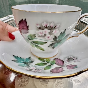 May include: White teacup and saucer set with gold trim. The cup and saucer feature a floral design with pink, purple, and green leaves and flowers. The teacup is on a silver tray.