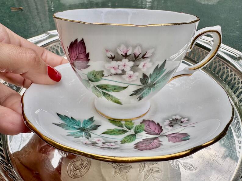 May include: White teacup and saucer set with gold trim. The cup and saucer feature a floral design with pink, purple, and green leaves and flowers. The teacup is on a silver tray.