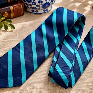 May include: A necktie featuring a repeating diagonal striped pattern in dark navy blue and bright turquoise. The fabric has a subtle ribbed texture, and the tie is laid out flat with the wider end angled toward the left and the narrow end folded.