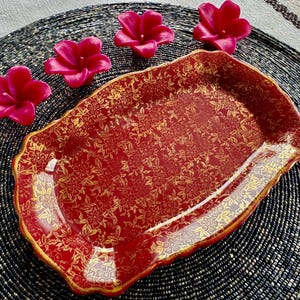 May include: A red and gold decorative serving tray with a floral pattern. The tray has a scalloped edge and is sitting on a black and white woven mat.