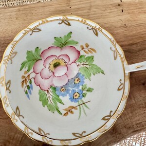 May include: A white teacup and saucer with a gold rim and a floral design. The design features pink and blue flowers with green leaves.