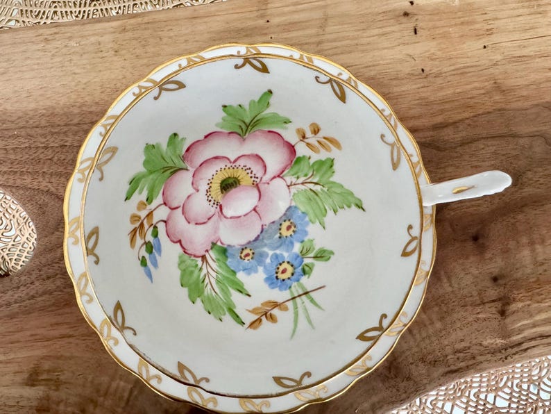 May include: A white teacup and saucer with a gold rim and a floral design. The design features pink and blue flowers with green leaves.