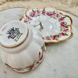 Puede incluir: Un juego de taza y platillo blancos con dise&ntilde;os florales de rosas rosas y ribetes dorados. La taza est&aacute; marcada "Salisbury Bone China England". El platillo tiene un borde festoneado. El juego est&aacute; sobre una bandeja de m&aacute;rmol.