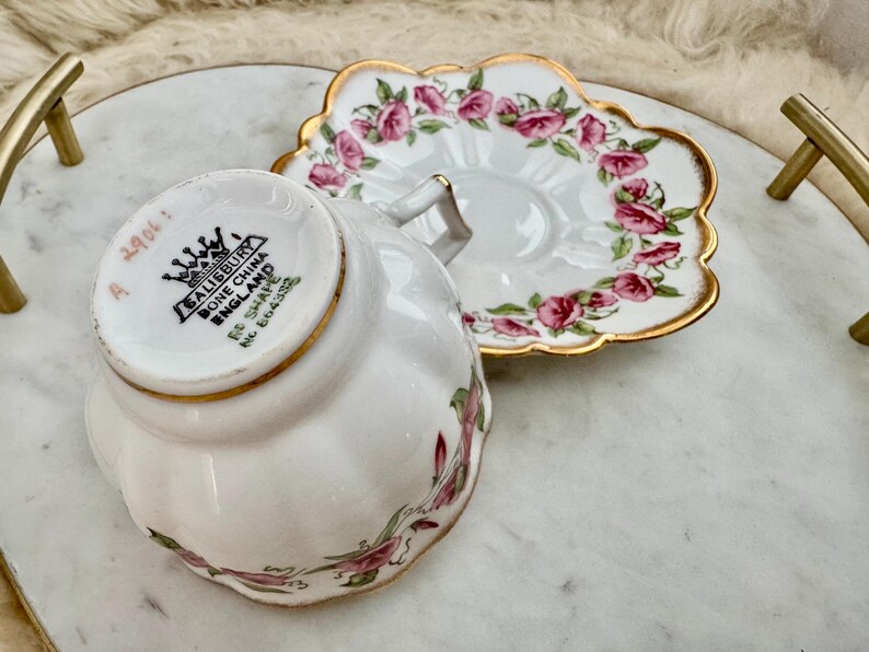 Puede incluir: Un juego de taza y platillo blancos con dise&ntilde;os florales de rosas rosas y ribetes dorados. La taza est&aacute; marcada "Salisbury Bone China England". El platillo tiene un borde festoneado. El juego est&aacute; sobre una bandeja de m&aacute;rmol.