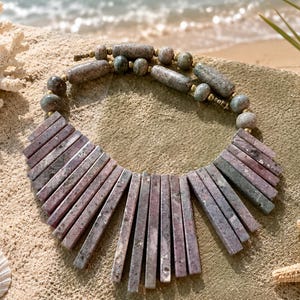 May include: A statement necklace featuring a series of rectangular, mauve-toned stone slabs arranged in a fan shape, connected to a strand of round and cylindrical speckled grey stone beads with small gold-toned metal spacers throughout.