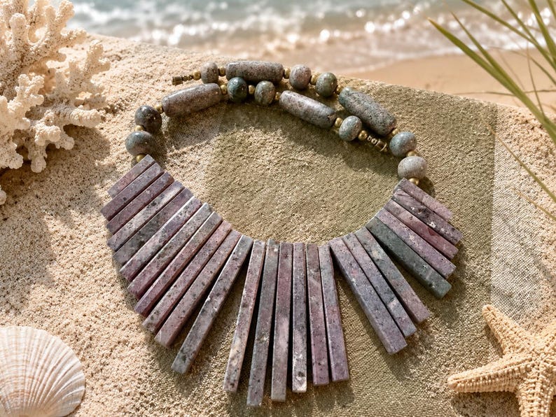 May include: A statement necklace featuring a series of rectangular, mauve-toned stone slabs arranged in a fan shape, connected to a strand of round and cylindrical speckled grey stone beads with small gold-toned metal spacers throughout.