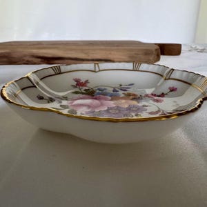 May include: A white, scalloped-edge dish with a floral pattern in pink, purple, and blue. The rim is trimmed with gold. The dish is likely made of porcelain or a similar material.