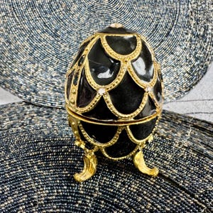 May include: A black and gold egg-shaped trinket box with a decorative design. The box has a hinged lid and sits on three gold feet.