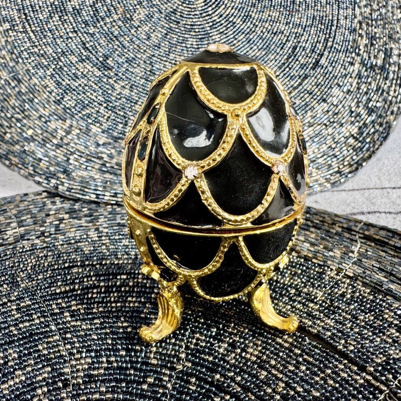 May include: A black and gold egg-shaped trinket box with a decorative design. The box has a hinged lid and sits on three gold feet.