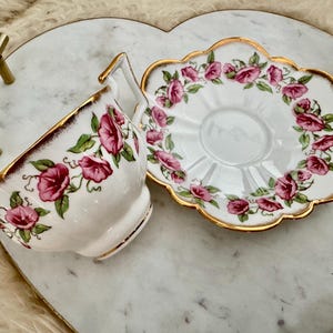 Puede incluir: Un juego de taza y platillo blancos con un dise&ntilde;o floral de flores rosas y hojas verdes. Los bordes est&aacute;n adornados con oro. El juego est&aacute; sobre una bandeja en forma de coraz&oacute;n con asas doradas.