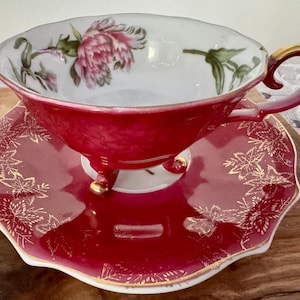 May include: A vintage teacup and saucer set. The teacup is a deep red with a gold-trimmed handle and footed base. The interior of the cup features a floral design with pink and green accents. The saucer is red with gold leaf patterns.