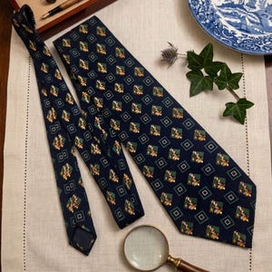 K&ouml;nnte beinhalten: Marineblaue Krawatte mit einem sich wiederholenden Muster aus gelben und gr&uuml;nen floralen Designs in gr&uuml;nen und schwarzen Quadraten. Die Krawatte wird auf einer cremefarbenen Oberfl&auml;che mit einer Lupe pr&auml;sentiert.