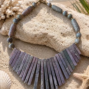 May include: A necklace featuring a strand of round and cylindrical beads in shades of gray, blue, and purple, leading to a central fan-shaped arrangement of vertical, rectangular stone segments in muted purple and green tones with a textured finish.
