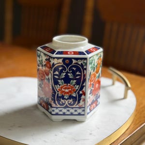 Peut inclure: Vase hexagonal en c&eacute;ramique avec une base blanche et un dessus &eacute;vas&eacute;. Le vase pr&eacute;sente un motif floral avec des fleurs rouges et oranges sur fond bleu. Le bord est orn&eacute; d'accents bleus et rouges.