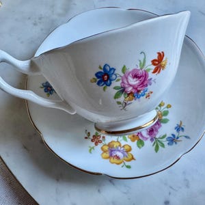 Puede incluir: Juego de taza y platillo blancos con un dise&ntilde;o floral con flores rosas, azules, naranjas y amarillas. La taza tiene un asa curva y un borde dorado, y el platillo tiene un borde festoneado con un ribete dorado.