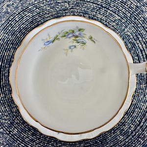 Pu&ograve; includere: Una tazza da t&egrave; e un piattino bianchi con un bordo dorato e un delicato disegno floreale con fiori blu.