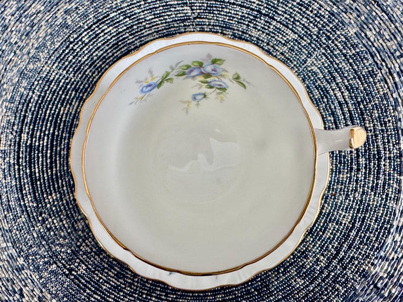 Pu&ograve; includere: Una tazza da t&egrave; e un piattino bianchi con un bordo dorato e un delicato disegno floreale con fiori blu.