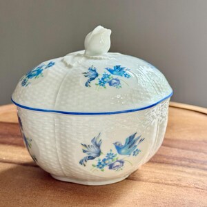 May include: A white ceramic lidded dish with a blue floral and bird design. The dish has a woven basket texture and a blue trim. The lid is topped with a white rose-shaped handle. The dish is sitting on a wooden surface.