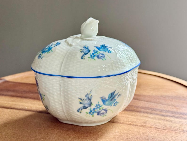 May include: A white ceramic lidded dish with a blue floral and bird design. The dish has a woven basket texture and a blue trim. The lid is topped with a white rose-shaped handle. The dish is sitting on a wooden surface.