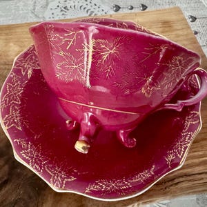 May include: A burgundy teacup and saucer set with gold leaf pattern detailing. The teacup has a handle and sits on a matching saucer. The set is elevated by small feet on the teacup. The edges of the saucer are scalloped.