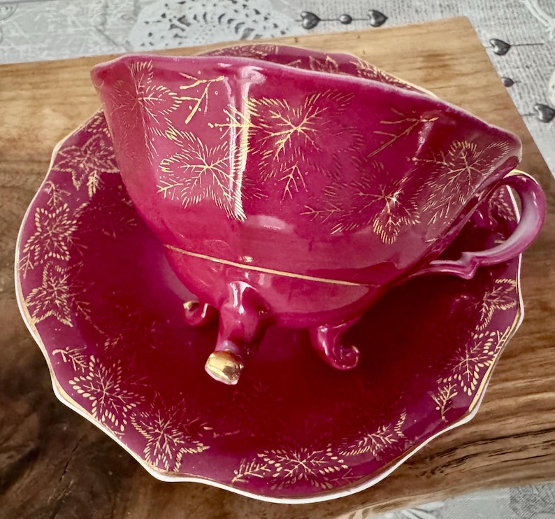 May include: A burgundy teacup and saucer set with gold leaf pattern detailing. The teacup has a handle and sits on a matching saucer. The set is elevated by small feet on the teacup. The edges of the saucer are scalloped.